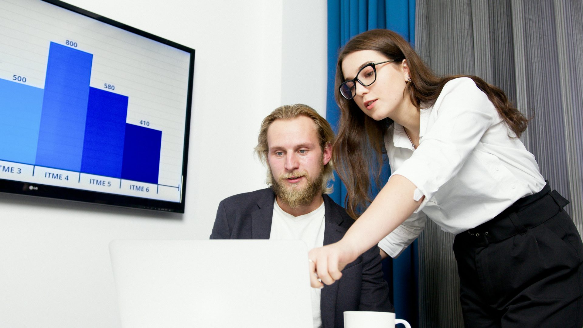 Two colleagues analyzing business data on a laptop with a presentation screen in the background.